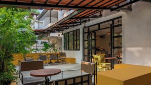 Un espace extérieur aménagé avec des tables et des chaises, entouré de plantes en pot et bordé d’un bâtiment doté de grandes fenêtres.