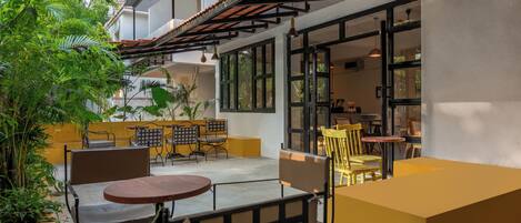 Un espace extérieur aménagé avec des tables et des chaises, entouré de plantes en pot et bordé d’un bâtiment doté de grandes fenêtres.