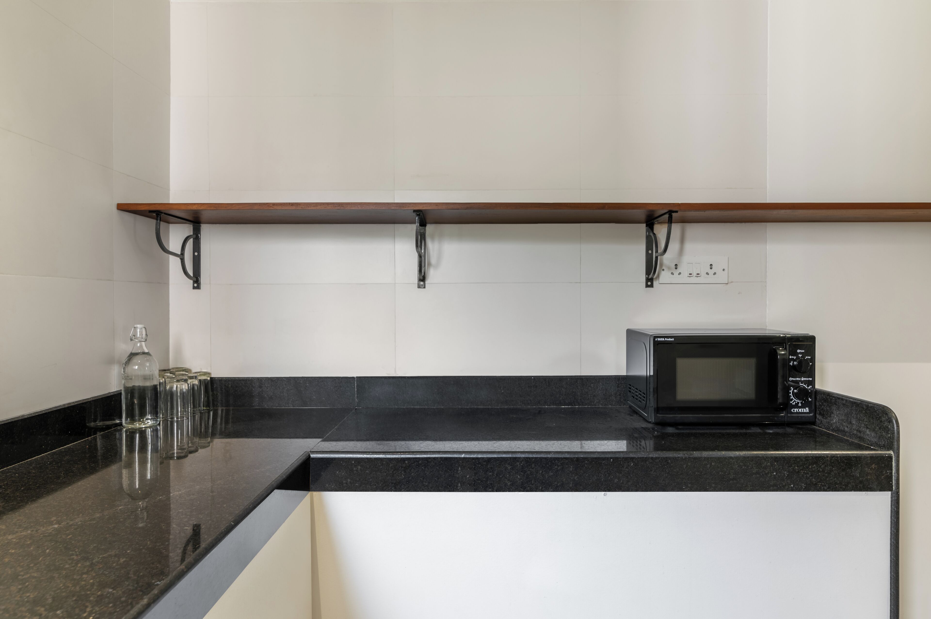 A kitchen with a black countertop, a microwave, a shelf, and a bottle.