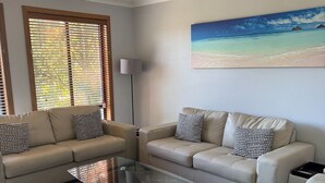 Een woonkamer met twee banken, een glazen salontafel, een plafondventilator en een wanddecoratie met een strandthema.