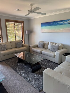 A living room with two sofas, a glass coffee table, a ceiling fan, and a beach mural on the wall.