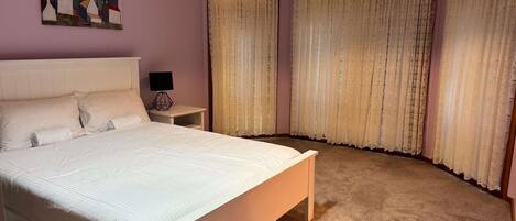 Une chambre à coucher avec un grand lit, une table de chevet avec une lampe et deux fenêtres ornées de rideaux légers.