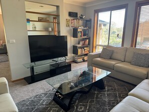 Ein Wohnzimmer mit einem Flachbildfernseher auf einem Glastisch, einem Bücherregal, einer Couch und einem Fenster mit Jalousien.