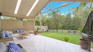 A covered outdoor seating area with a sofa and coffee table, surrounded by a garden and a view of trees. - Relax at the Park by Book Jervis Bay (Sanctuary Point)