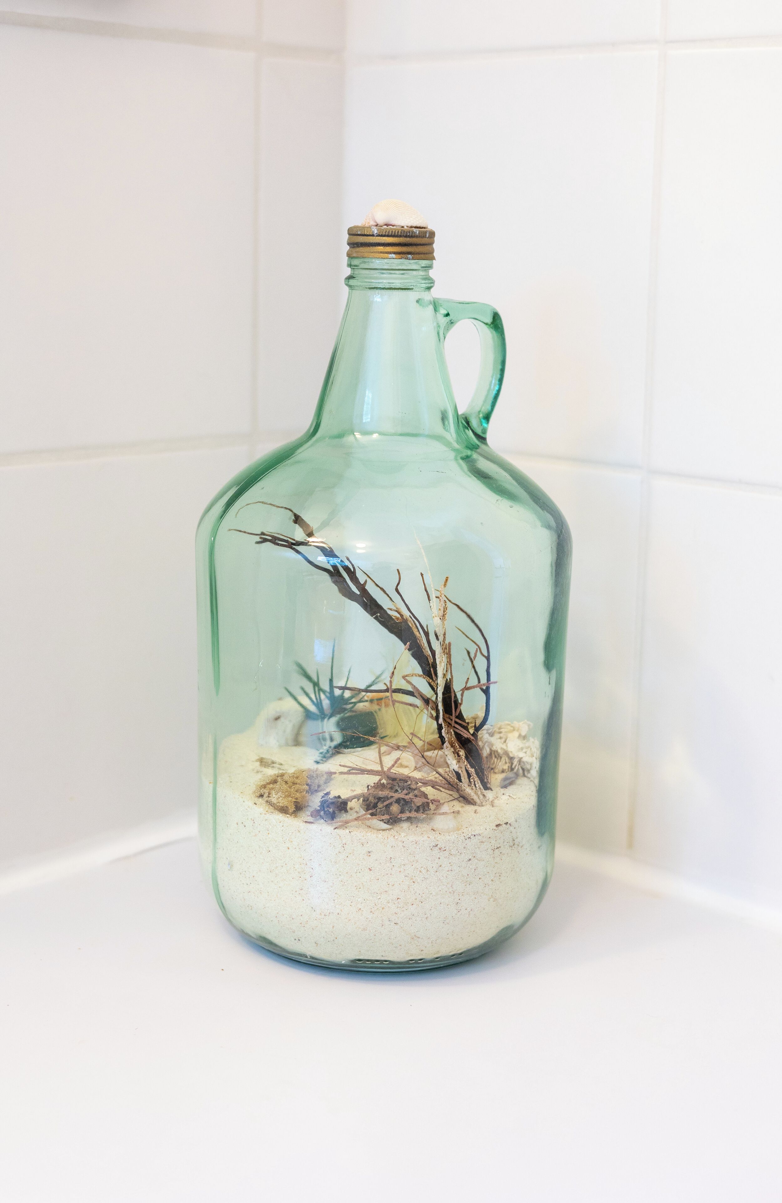 A clear glass bottle with a handle, containing a small arrangement of branches and pebbles.