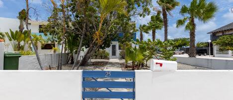 A gated entrance with a blue gate labeled '25', surrounded by lush tropical vegetation and a white wall.