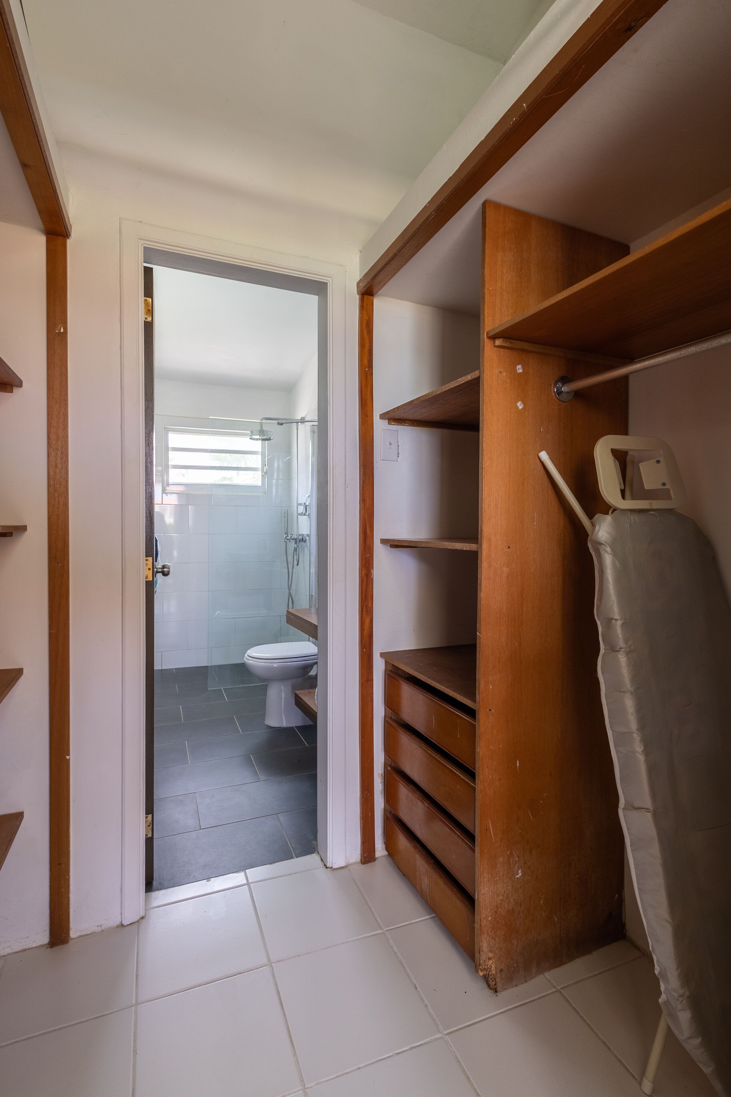 A bathroom with a toilet, a shower, and a storage closet with shelves and a hanging rack.