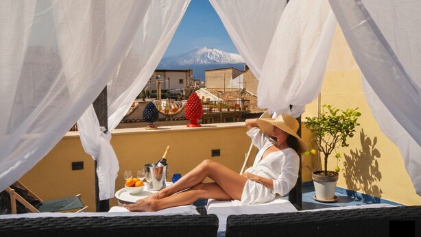 A person relaxing on a balcony with a view of a mountain, a table with a bottle and glasses, and a potted plant.