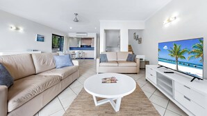 Ein modernes Wohnzimmer mit einem beigefarbenen Sofa, einem weißen Couchtisch und einem Fernseher, der eine Strandansicht zeigt.
