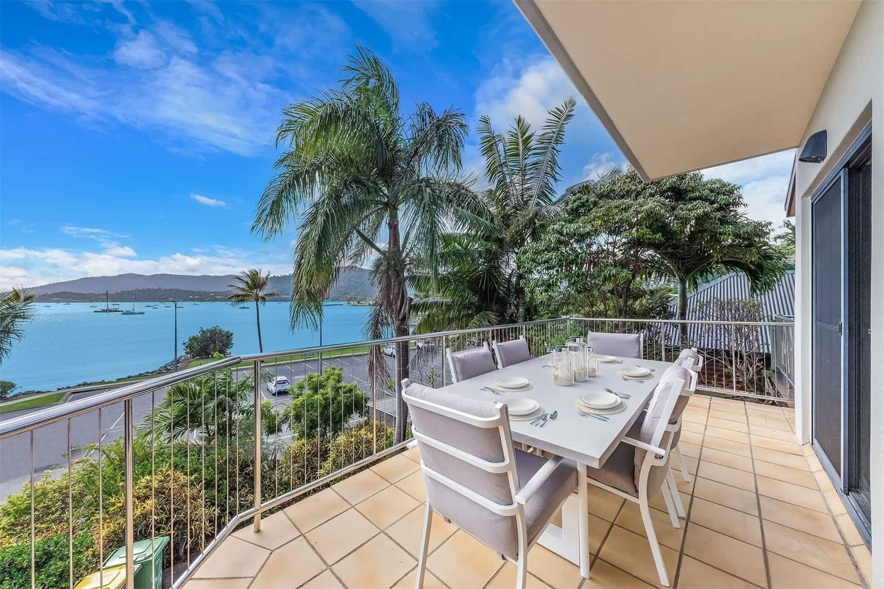 A balcony with a table set for dining, overlooking a body of water with boats and palm trees.