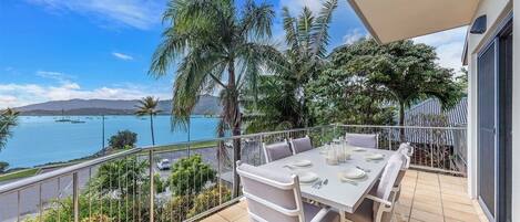 A balcony with a table set for dining, overlooking a body of water with boats and palm trees.