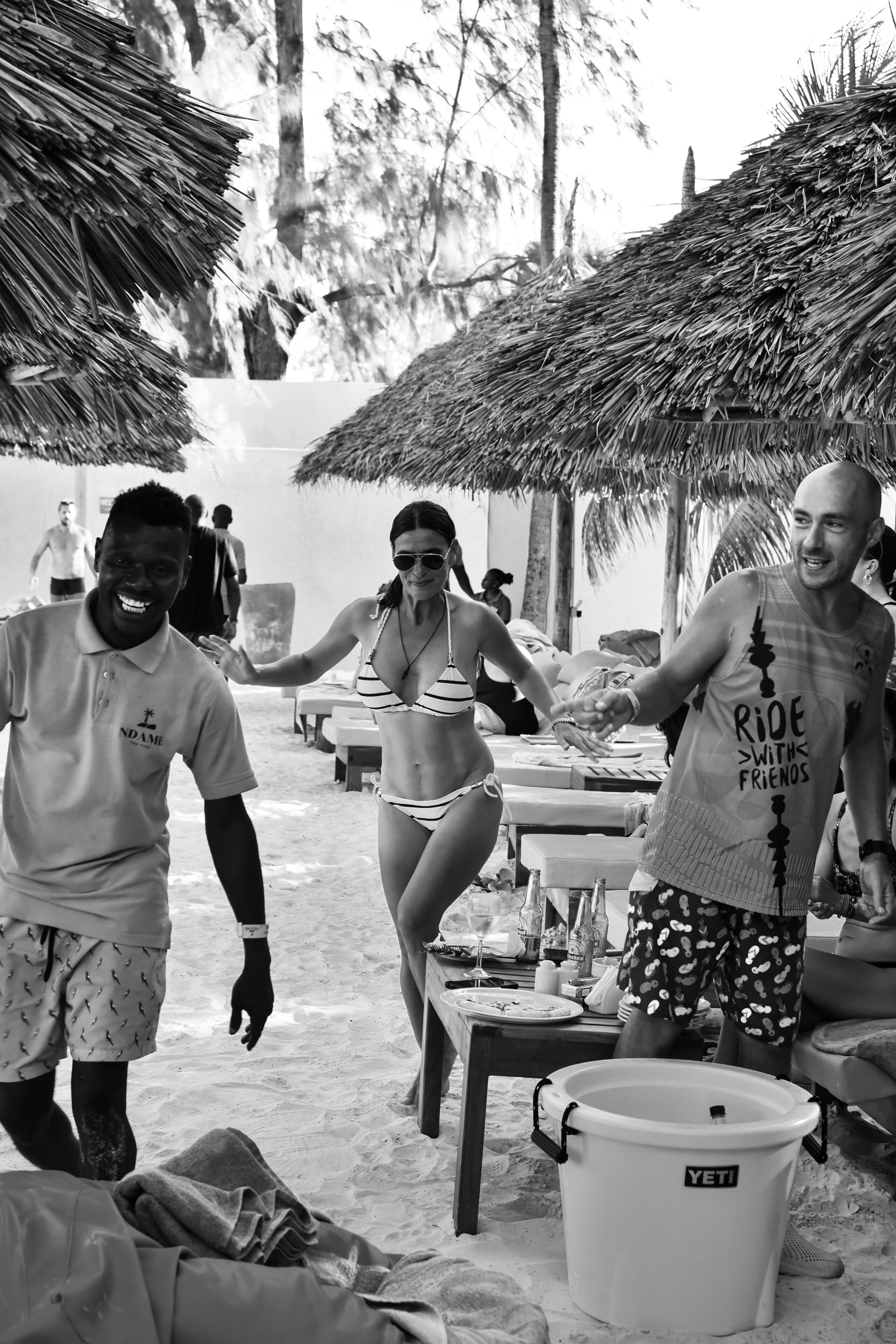Escena frente al mar con sombrillas de paja, una mesa de comedor con comida y bebidas, y un cubo con la etiqueta "YETI".