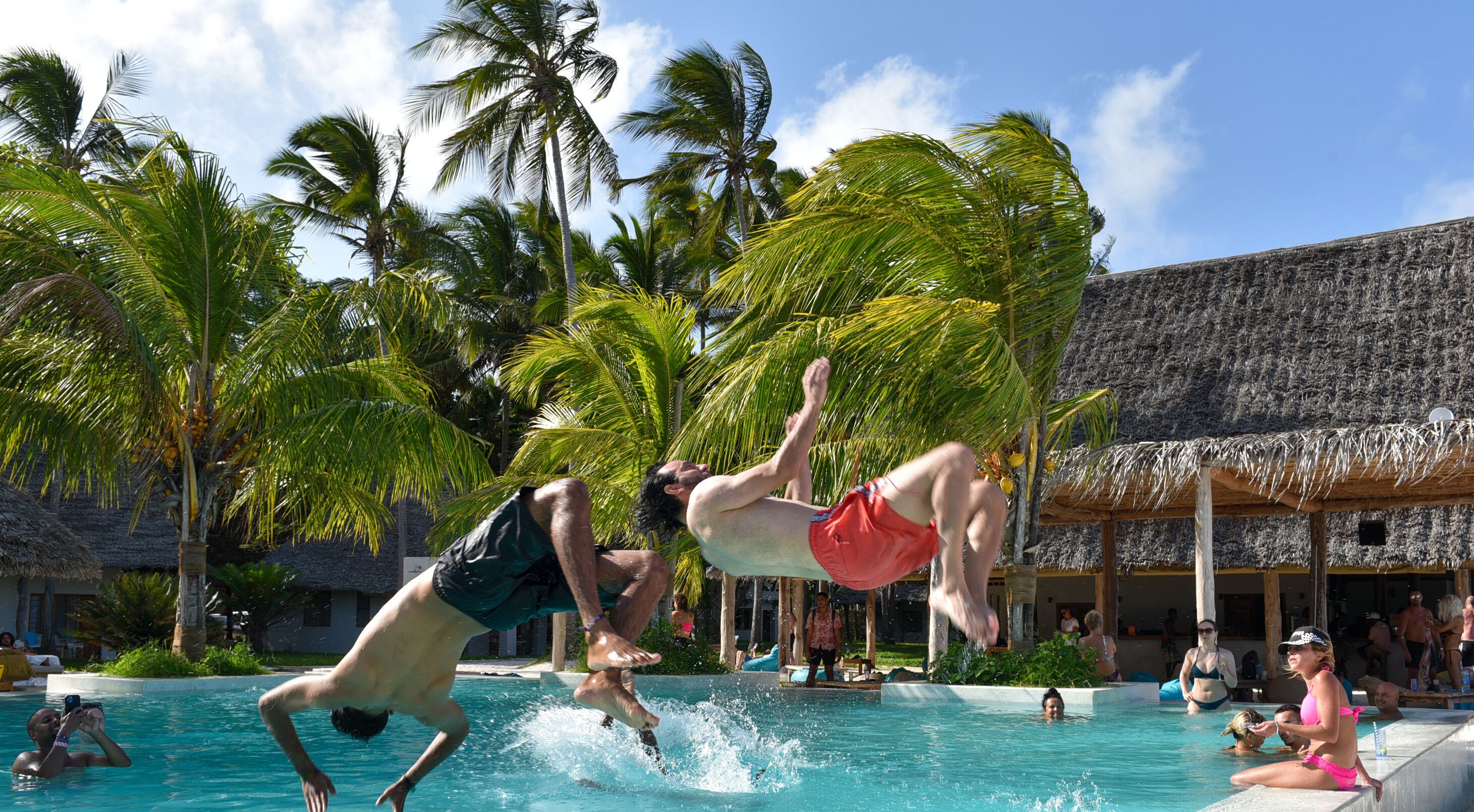 A pool area with palm trees and a thatched-roof building in the background.