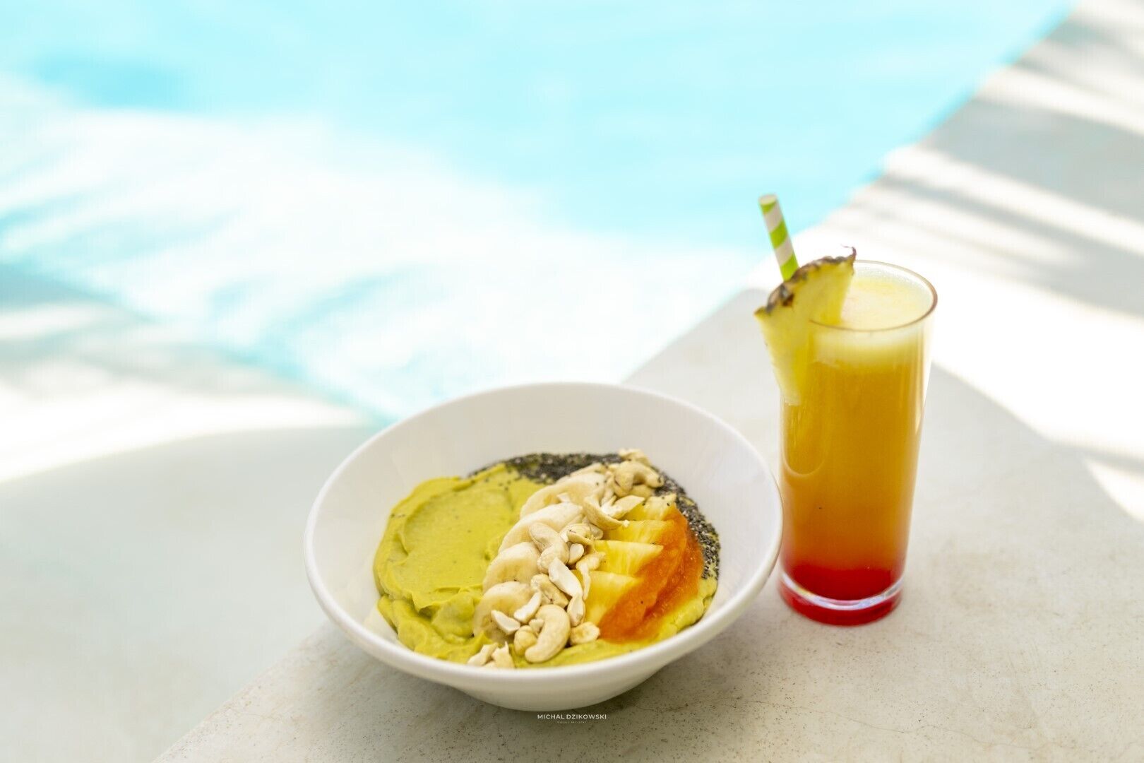 A bowl of food with cashews and pineapple on a white plate next to a glass of orange juice with a straw.
