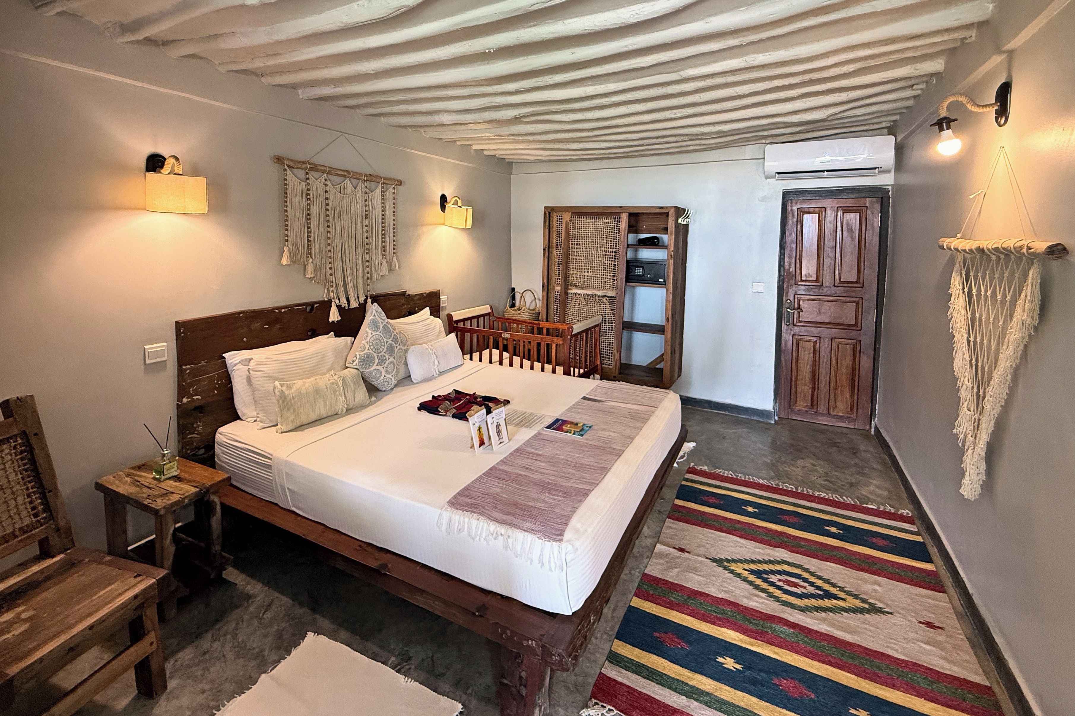 A bedroom with a large bed, wooden headboard, bedside tables, a nightstand, a wooden wardrobe, a door, and a macrame wall hanging.