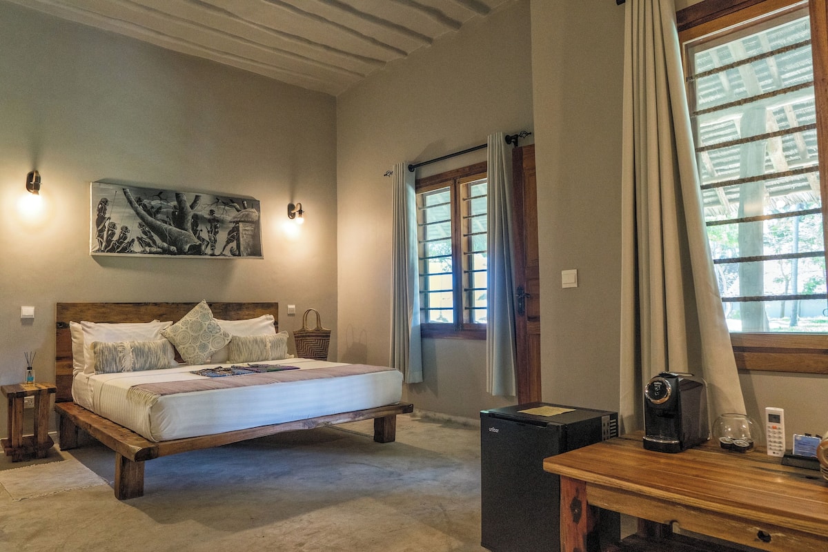 A bedroom with a bed, a bedside table, a coffee maker, and a window with curtains.