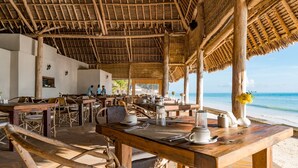 An open-air dining area with wooden tables and chairs, a thatched roof, and a view of the beach. - Ndame Paje Hotel (Paje)