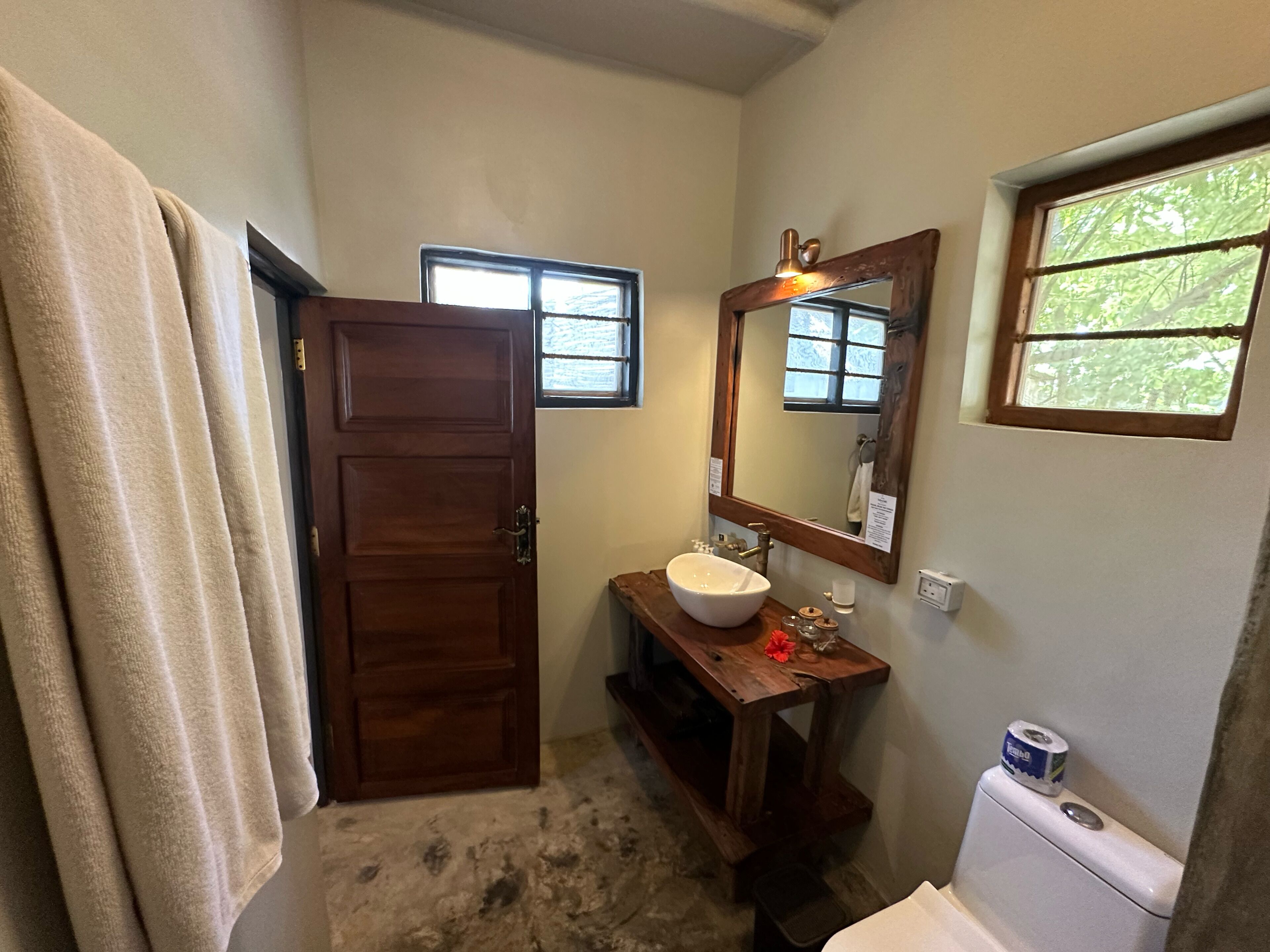 Un baño con puerta de madera, un lavabo con espejo, una ventana y un inodoro.