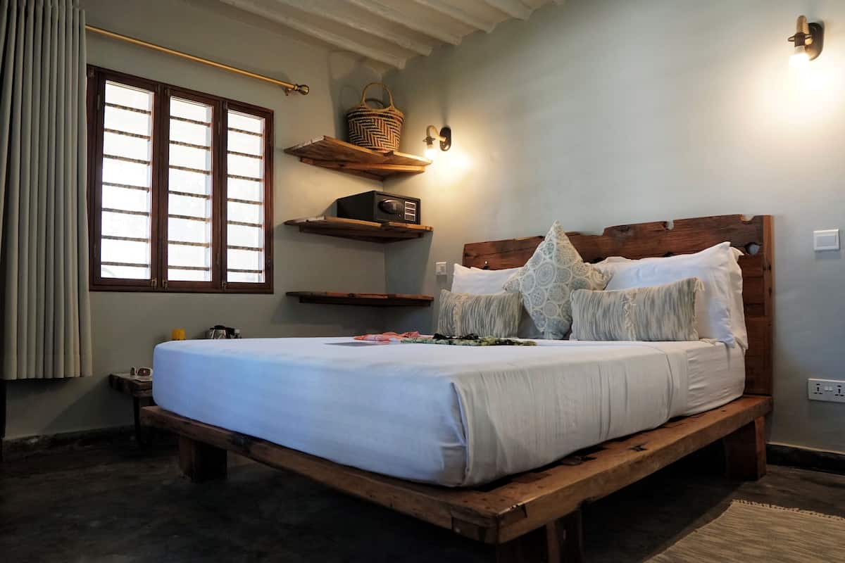 A bedroom with a wooden bed, a window with blinds, a bedside table, and a wall-mounted shelf with a basket.