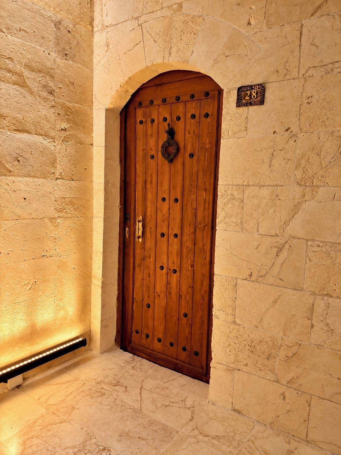 Una puerta de madera con un aldabón de metal, ubicada en un muro de piedra con una placa numérica.