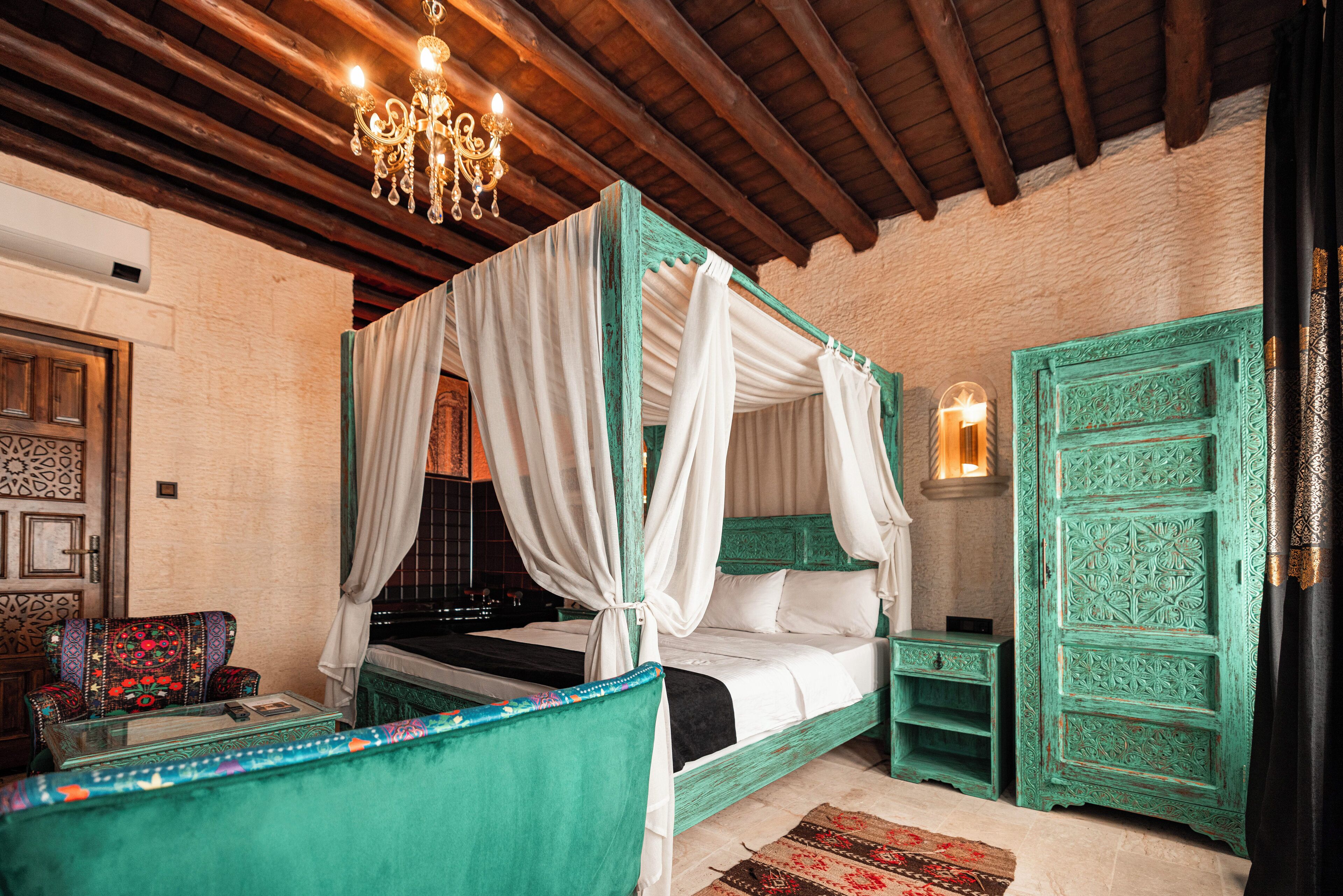 Una cama con dosel con ropa blanca, un sillón tapizado en verde, una mesita con un libro y un control remoto, y un panel decorativo en la pared.