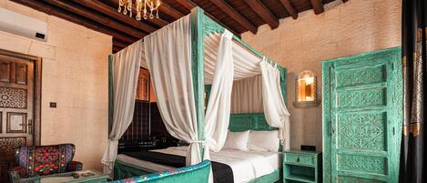 Una cama con dosel con ropa blanca, un sillón tapizado en verde, una mesita con un libro y un control remoto, y un panel decorativo en la pared.