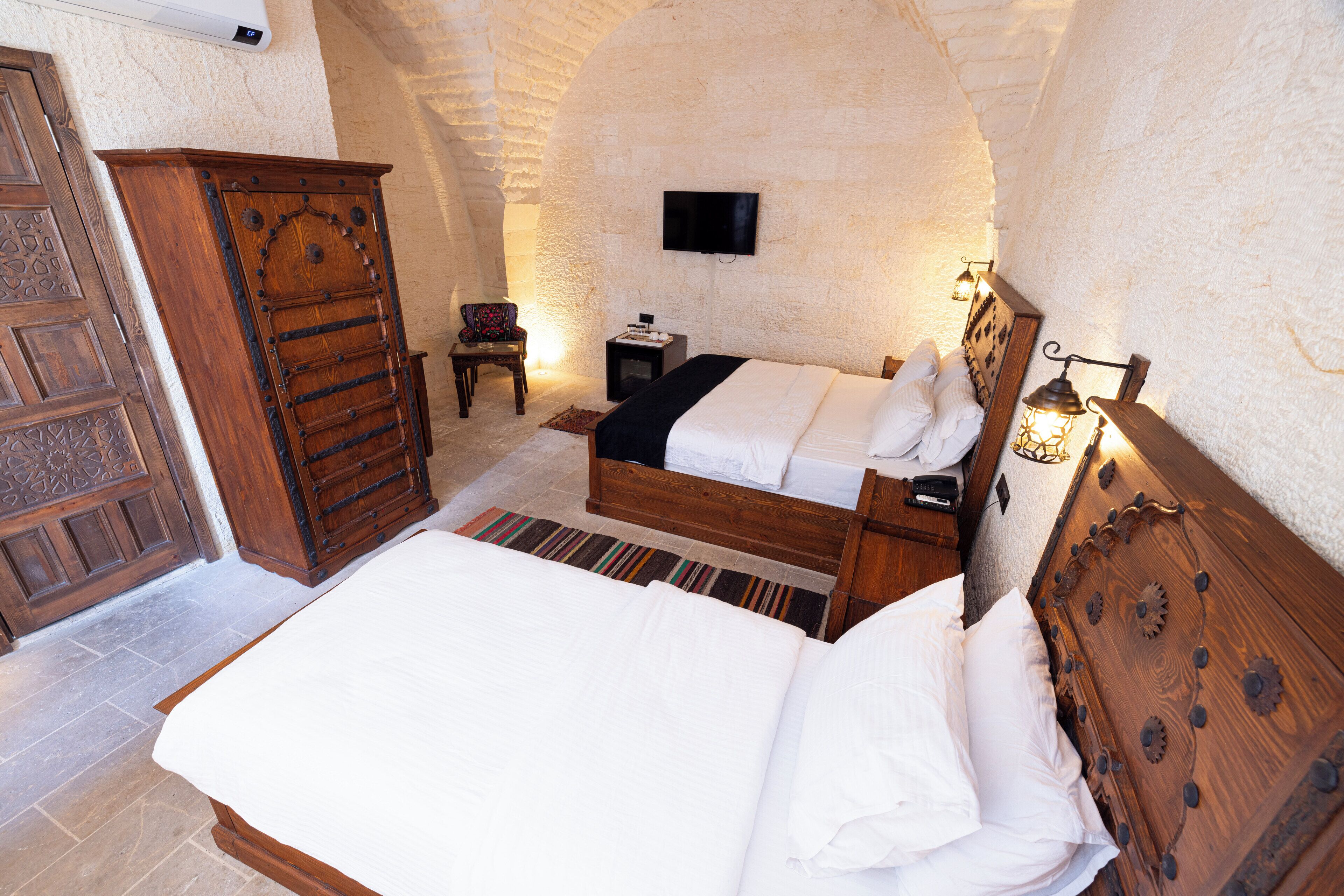 Quarto com cama de madeira, guarda-roupa, televisão na parede e uma mesinha com abajur.