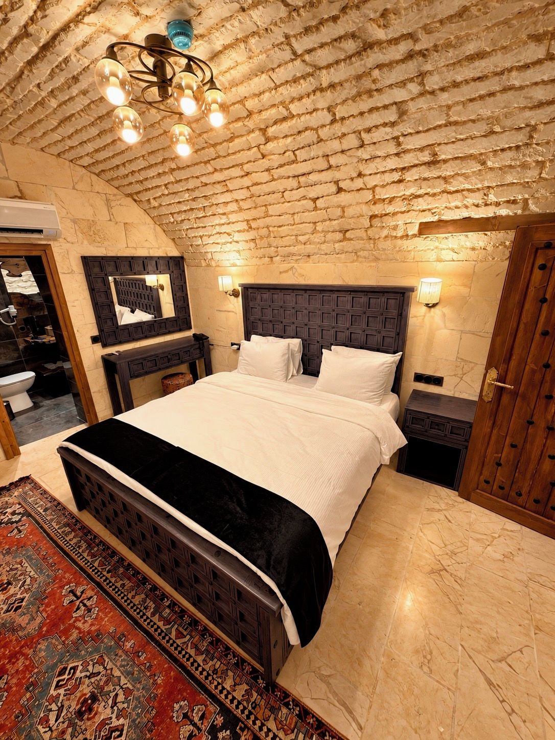 A bedroom with a brick ceiling, a large bed with a dark headboard, bedside tables, and a patterned rug.