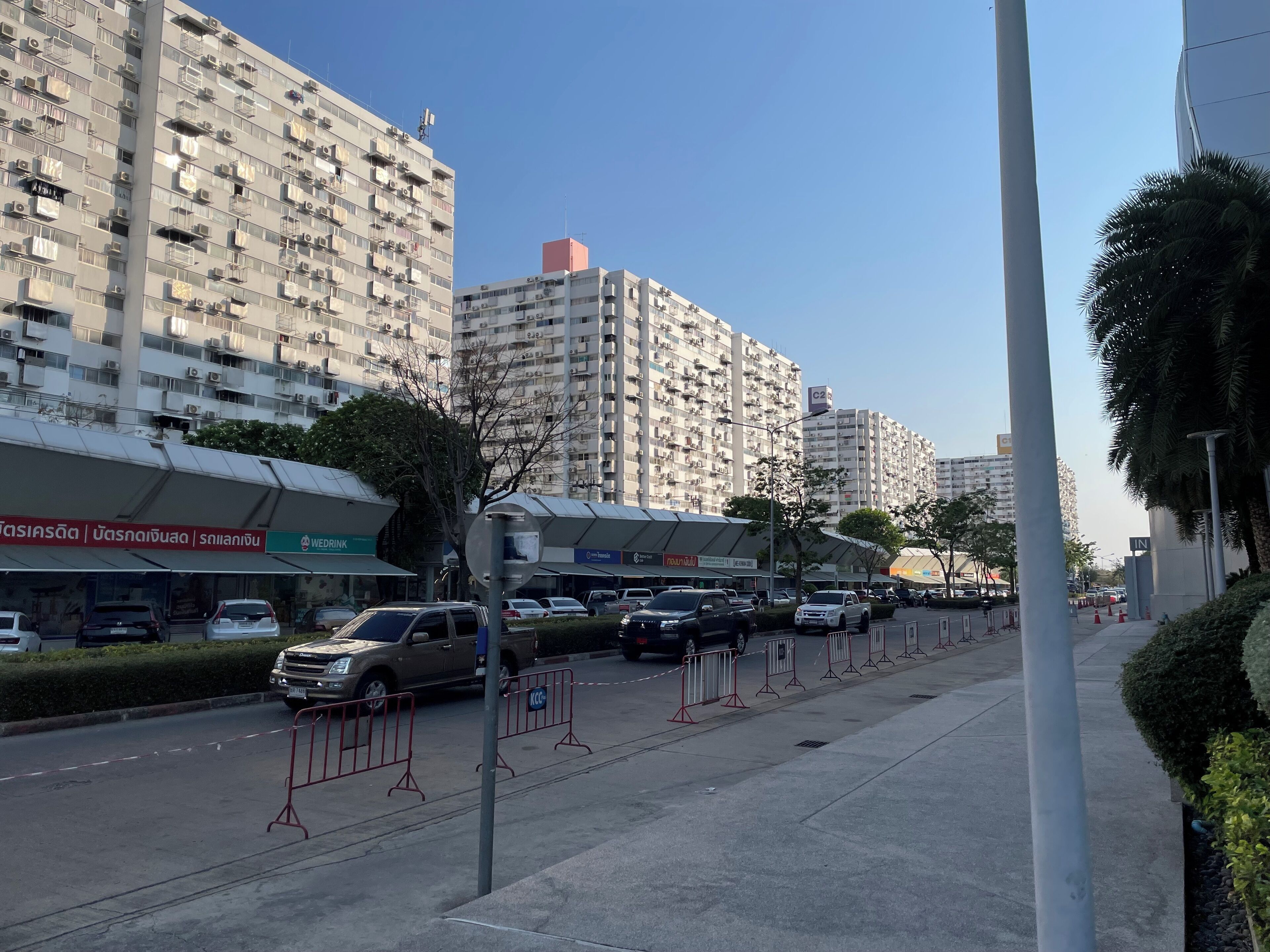 Una veduta di strada con edifici residenziali alti, auto parcheggiate e un marciapiede delimitato da barriere rosse.