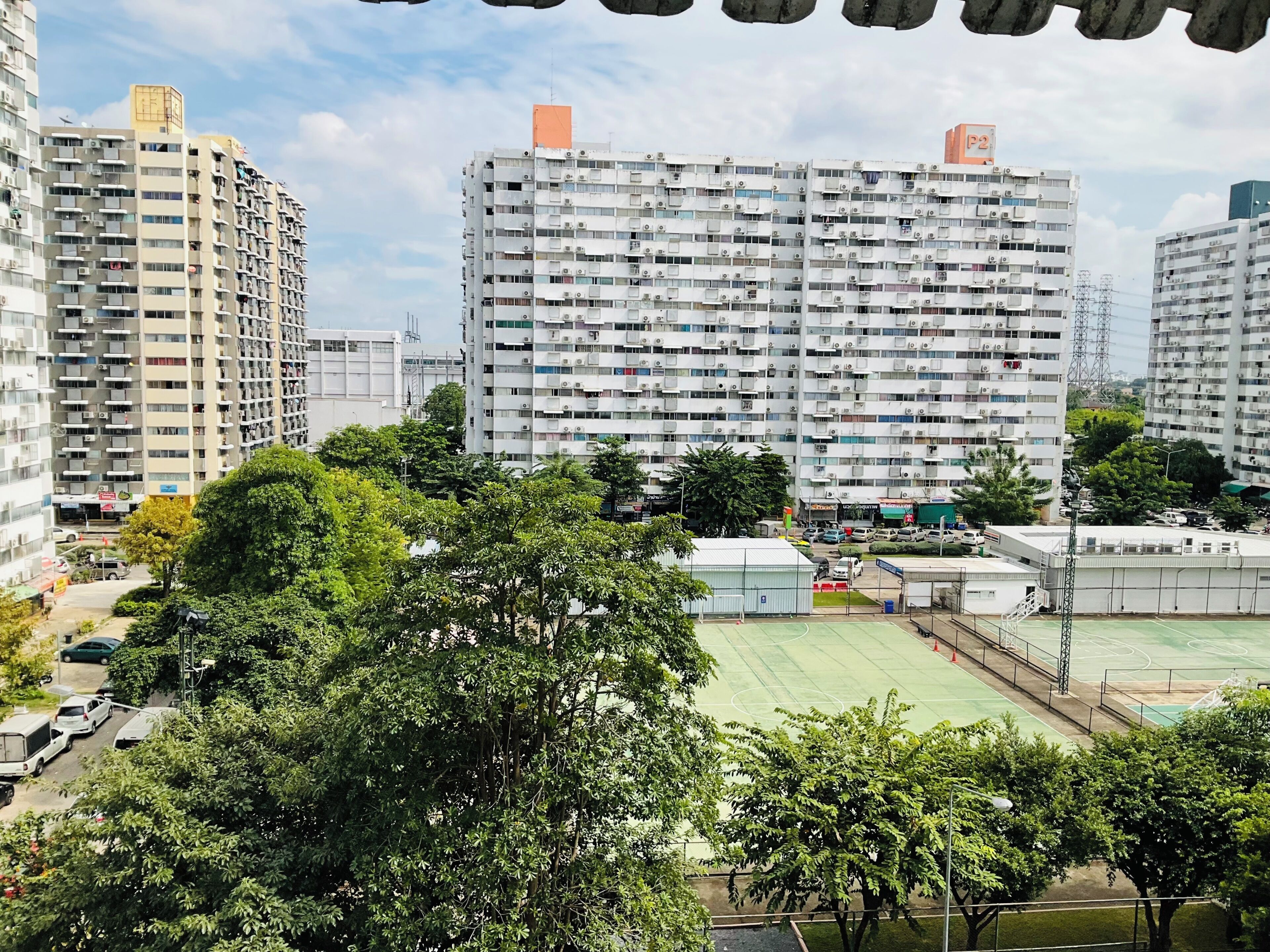 A high-rise residential building with multiple balconies, a sports court, and surrounding greenery.