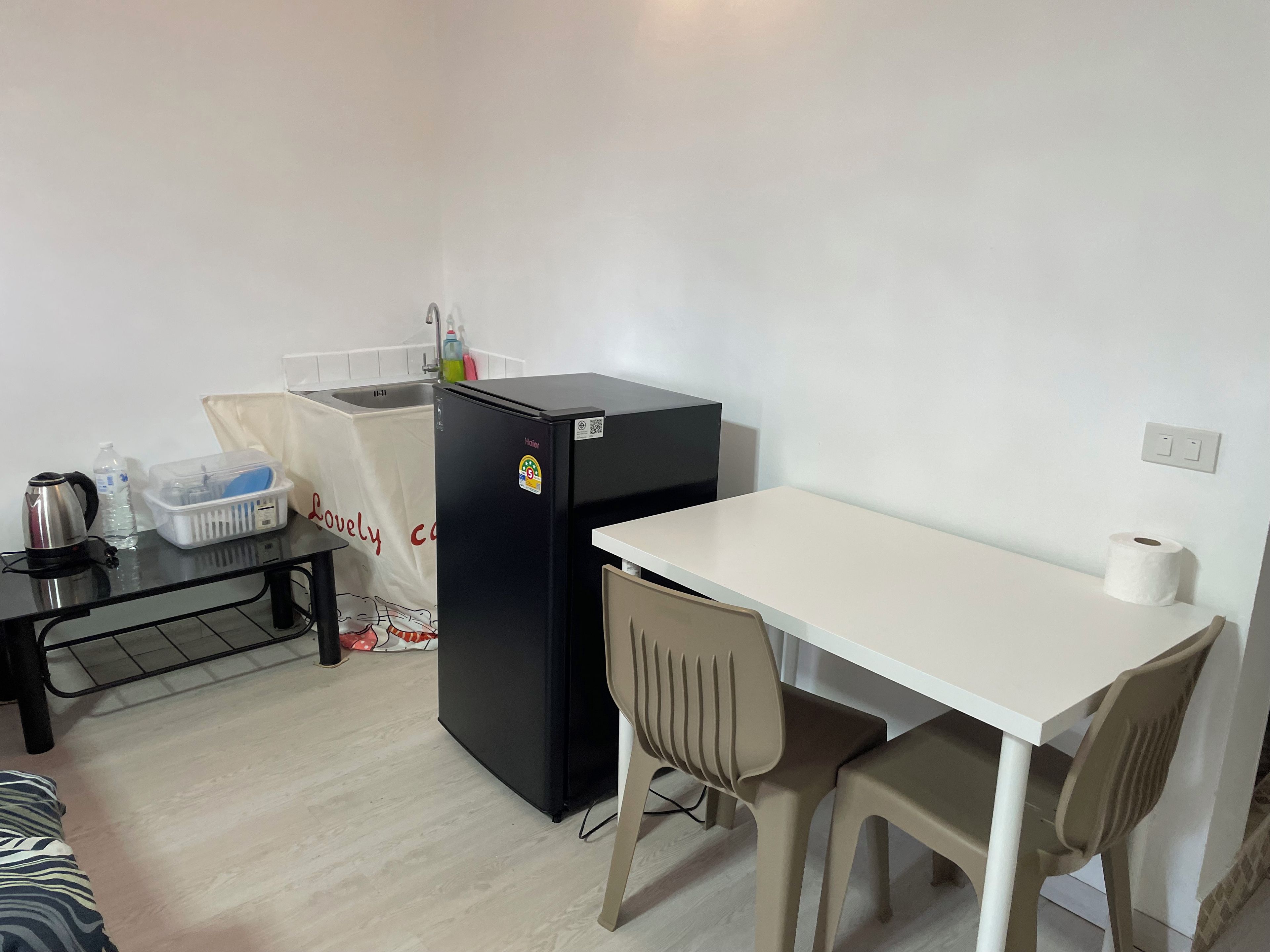 Uma cozinha compacta equipada com um frigorífico, uma pequena mesa, uma cadeira, uma chaleira e um recipiente de plástico.
