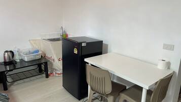 Uma cozinha compacta equipada com um frigorífico, uma pequena mesa, uma cadeira, uma chaleira e um recipiente de plástico.