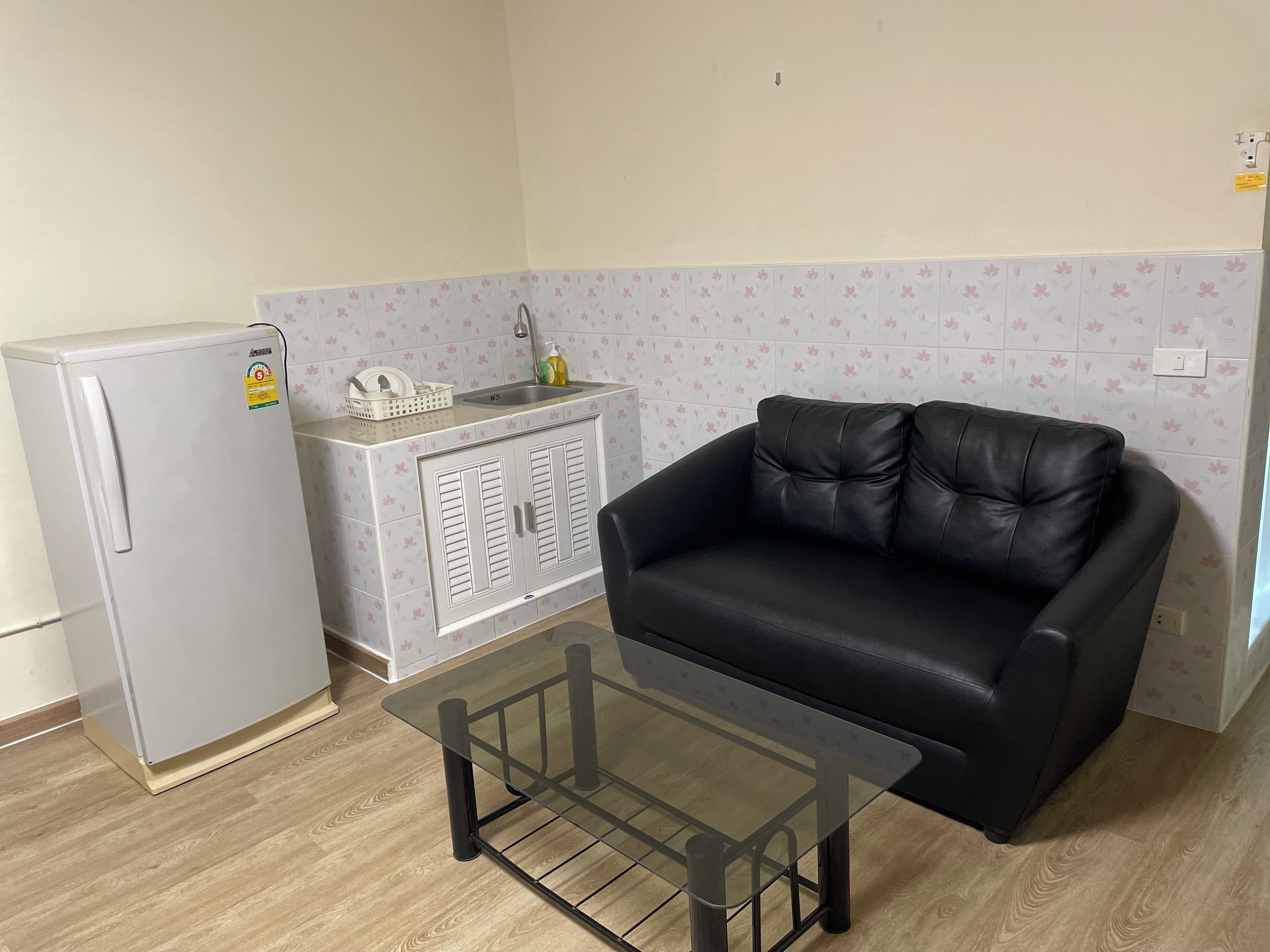 A compact kitchenette with a refrigerator, a small sink, and a cabinet. There's a black leather sofa and a glass-top coffee table in the living area.