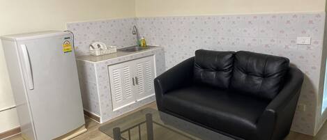 A compact kitchenette with a refrigerator, a small sink, and a cabinet. There's a black leather sofa and a glass-top coffee table in the living area.