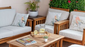 Un espace extérieur accueillant, aménagé avec des meubles en bois et des coussins, une petite table avec un magazine, une carafe de limonade et une plante en pot.