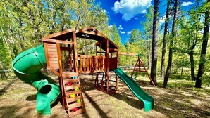 Un parque infantil con tobogán, pared de escalada y columpios en un área boscosa.