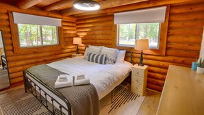 Un dormitorio tipo cabaña de madera con una cama, mesitas de noche, un escritorio y dos ventanas con persianas.