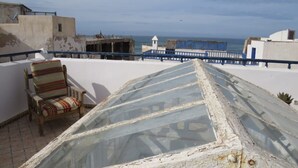 A rooftop terrace with a view of the sea, a striped chair, and a glass greenhouse structure. - Riad El Baze (Essaouira)