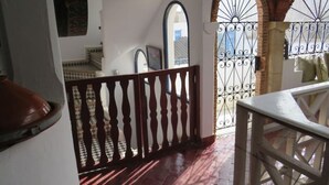 A hallway with a wooden railing, a tiled floor, and a view through an arched window. - Riad El Baze (Essaouira)