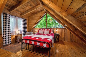 A wooden cabin bedroom with a sloped ceiling, a bed with a red and white quilt, a window with a view of trees, and a small bedside table with a lamp.
