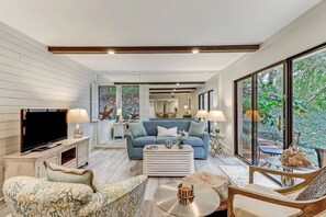 Un salon moderne avec un canapé, une table basse et un téléviseur. Une grande fenêtre offre une vue sur la verdure.