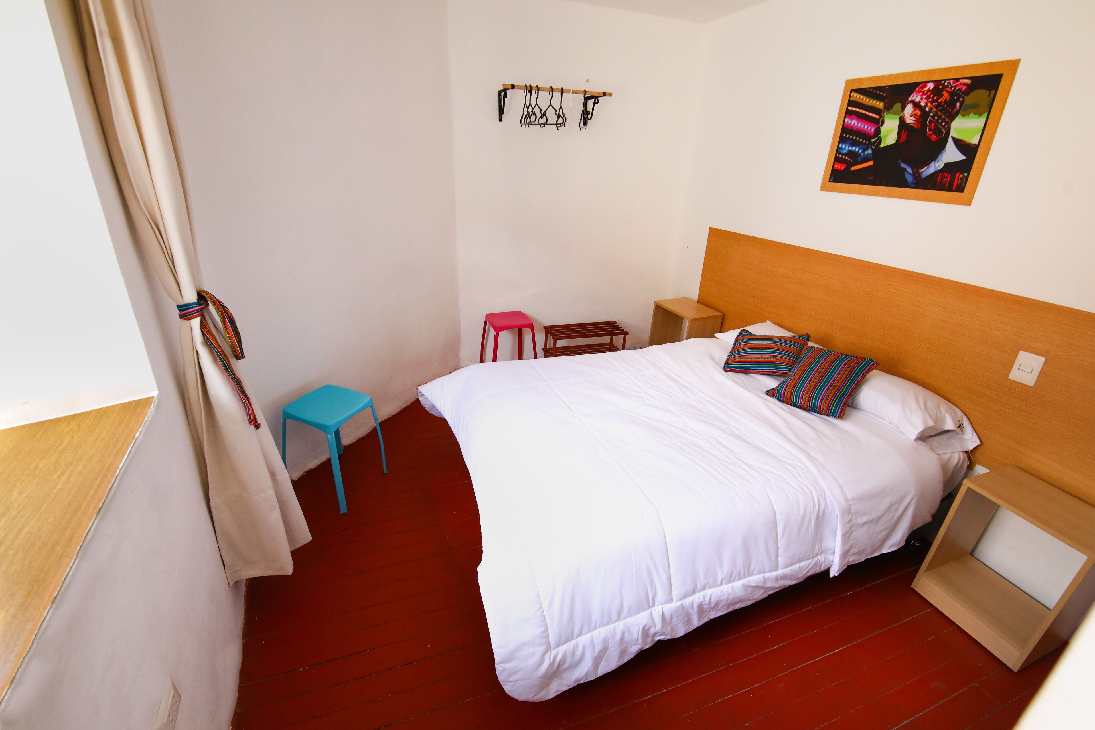 Un dormitorio con cabecera de madera, una cama blanca, un taburete rojo, una mesita y una lámpara fijada a la pared.