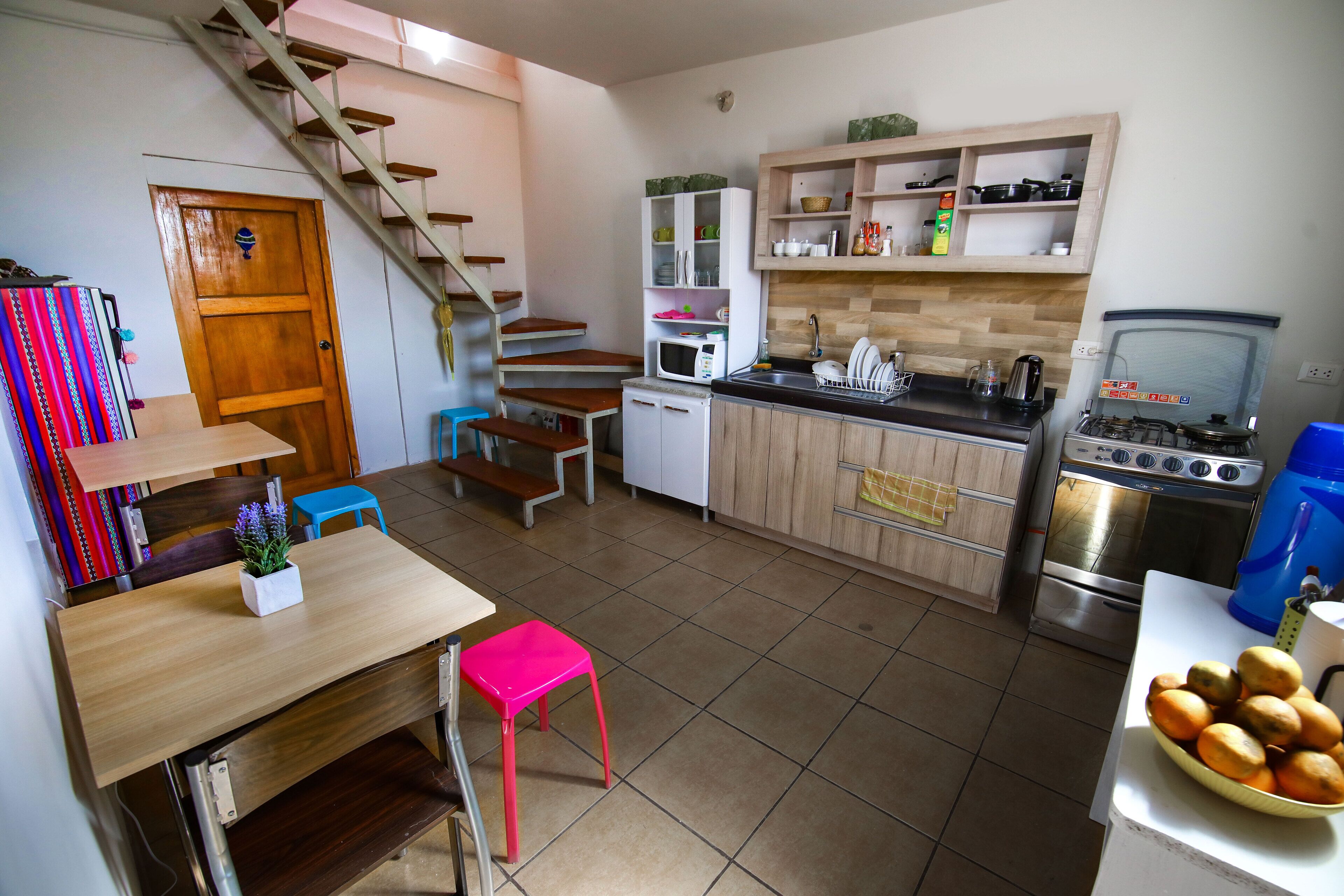 A compact kitchen with a wooden table, a pink stool, a microwave, a sink, and a stove.