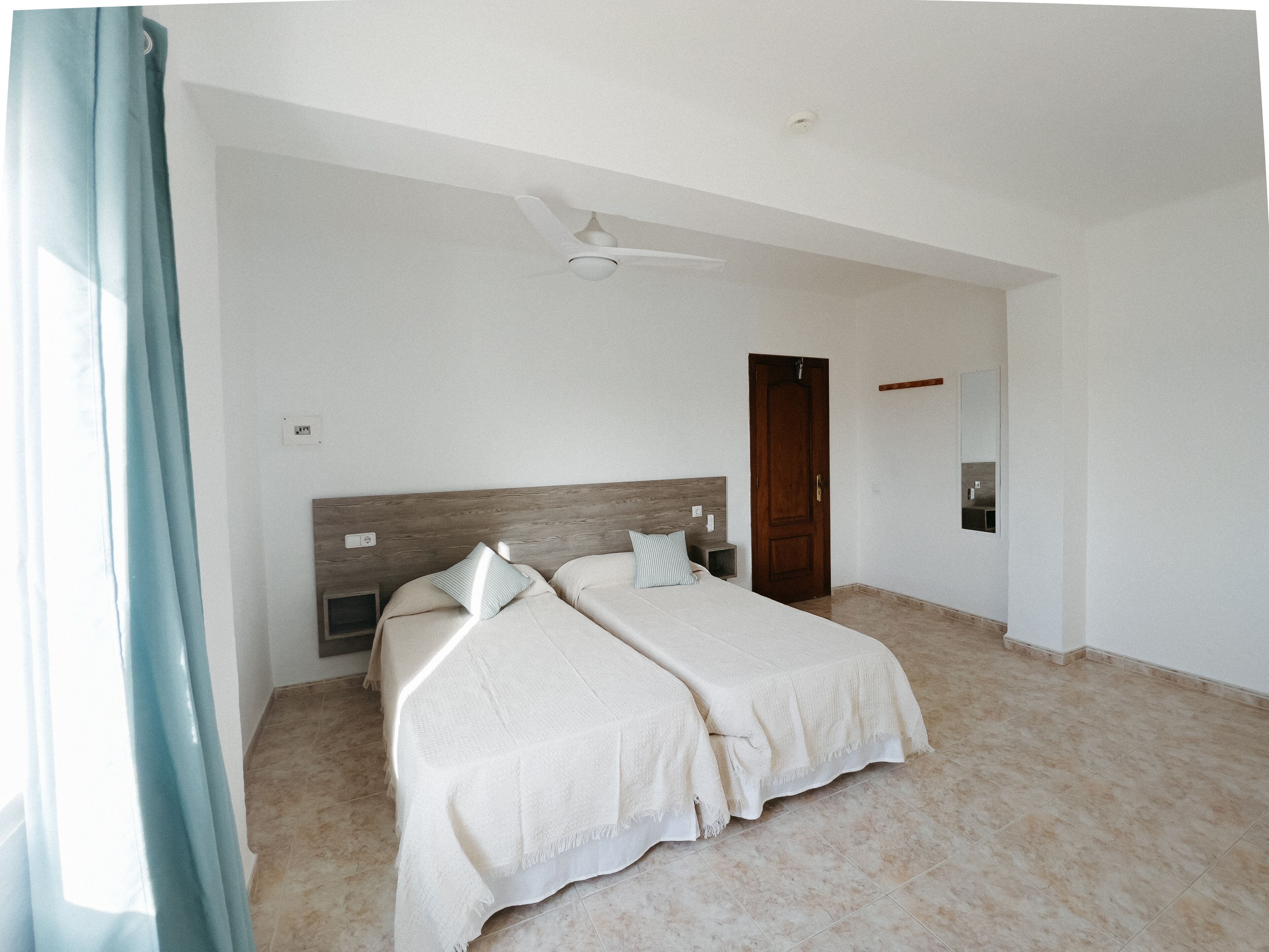 Un dormitorio con dos camas, un ventilador de techo, un cabecero de madera, una puerta y un espejo en la pared.