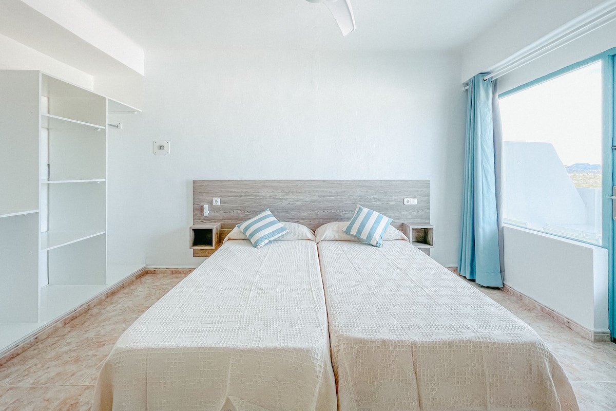 Un dormitorio con una cama grande, ventilador de techo, ventana con cortinas y vistas al exterior.