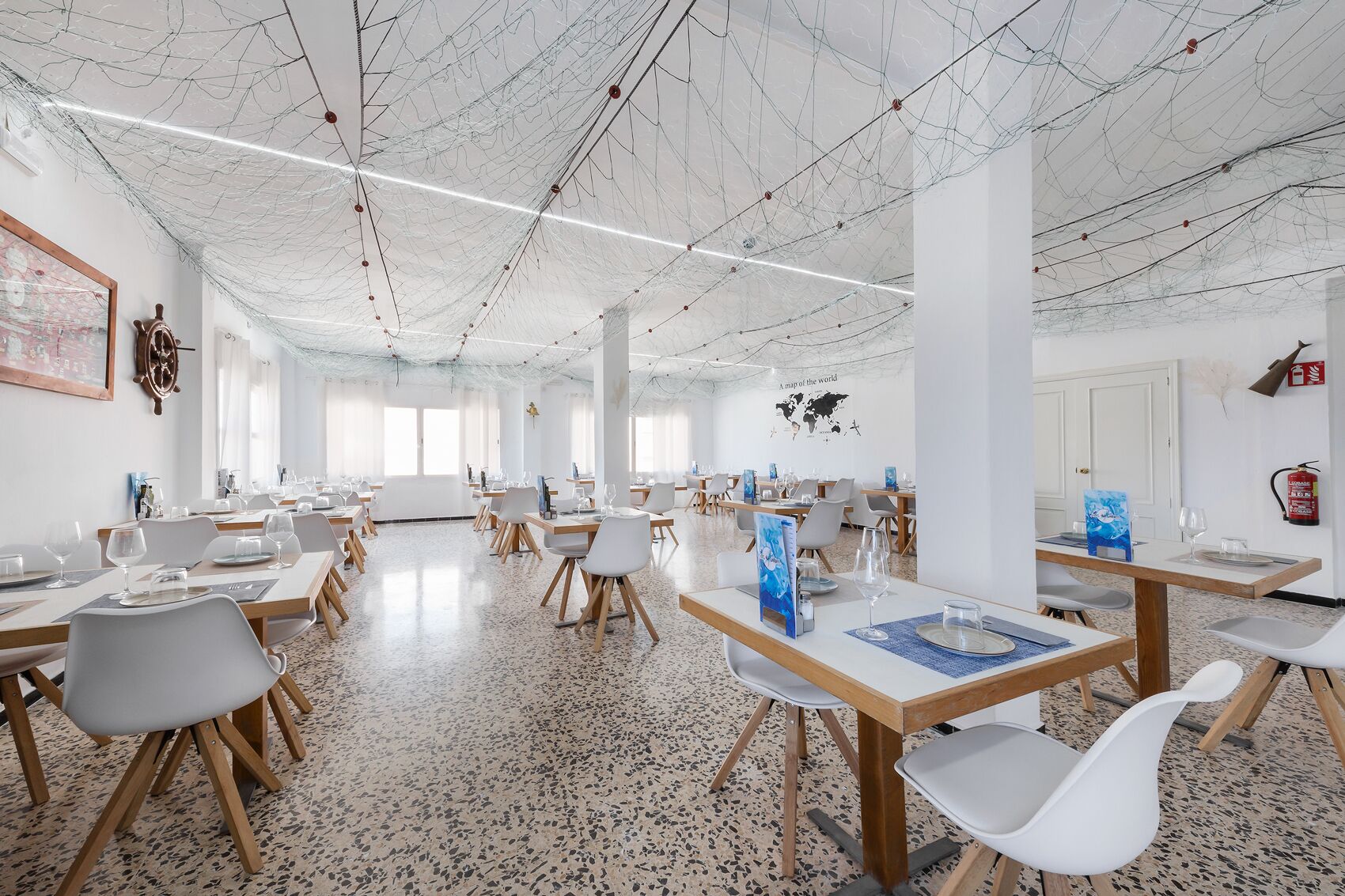 A spacious dining area with multiple tables and chairs, a high ceiling with exposed wiring, and a world map on the wall.