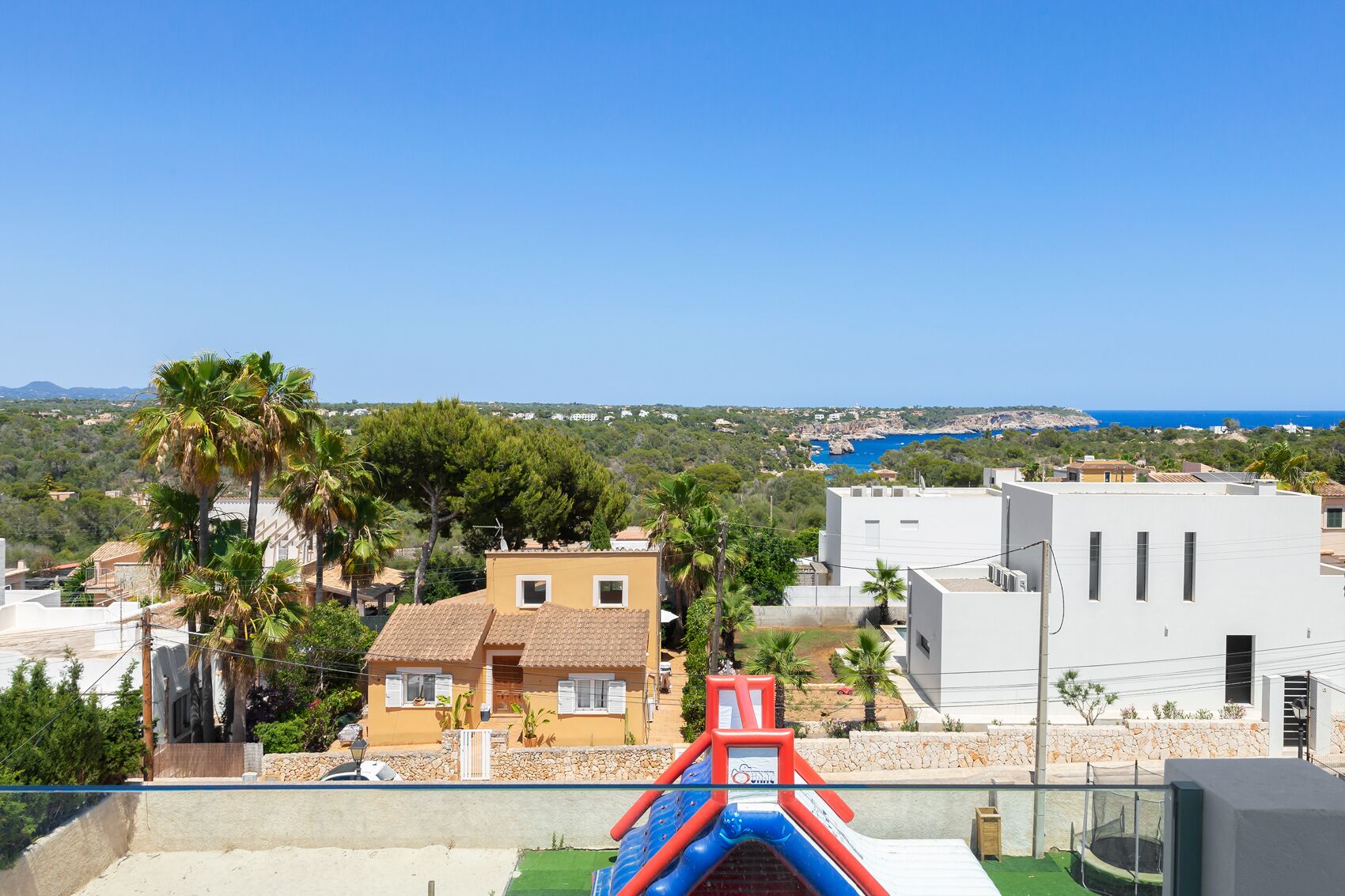 A residential area with a view of the sea, featuring houses with tiled roofs, palm trees, and a clear blue sky.