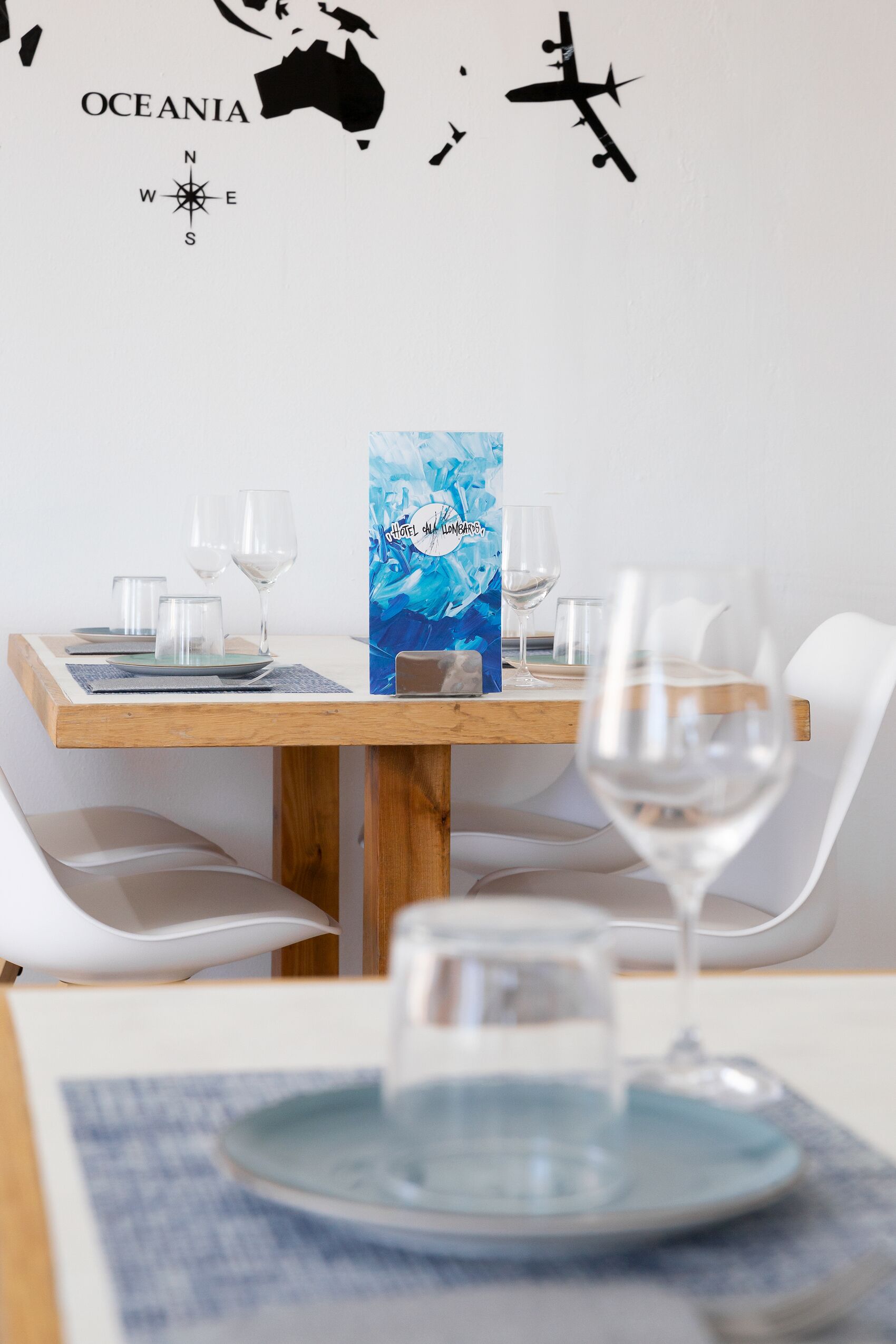 Una mesa de comedor dispuesta con copas de vino y platos, un atril para el menú, y una pared decorada con una brújula y una silueta de avión.