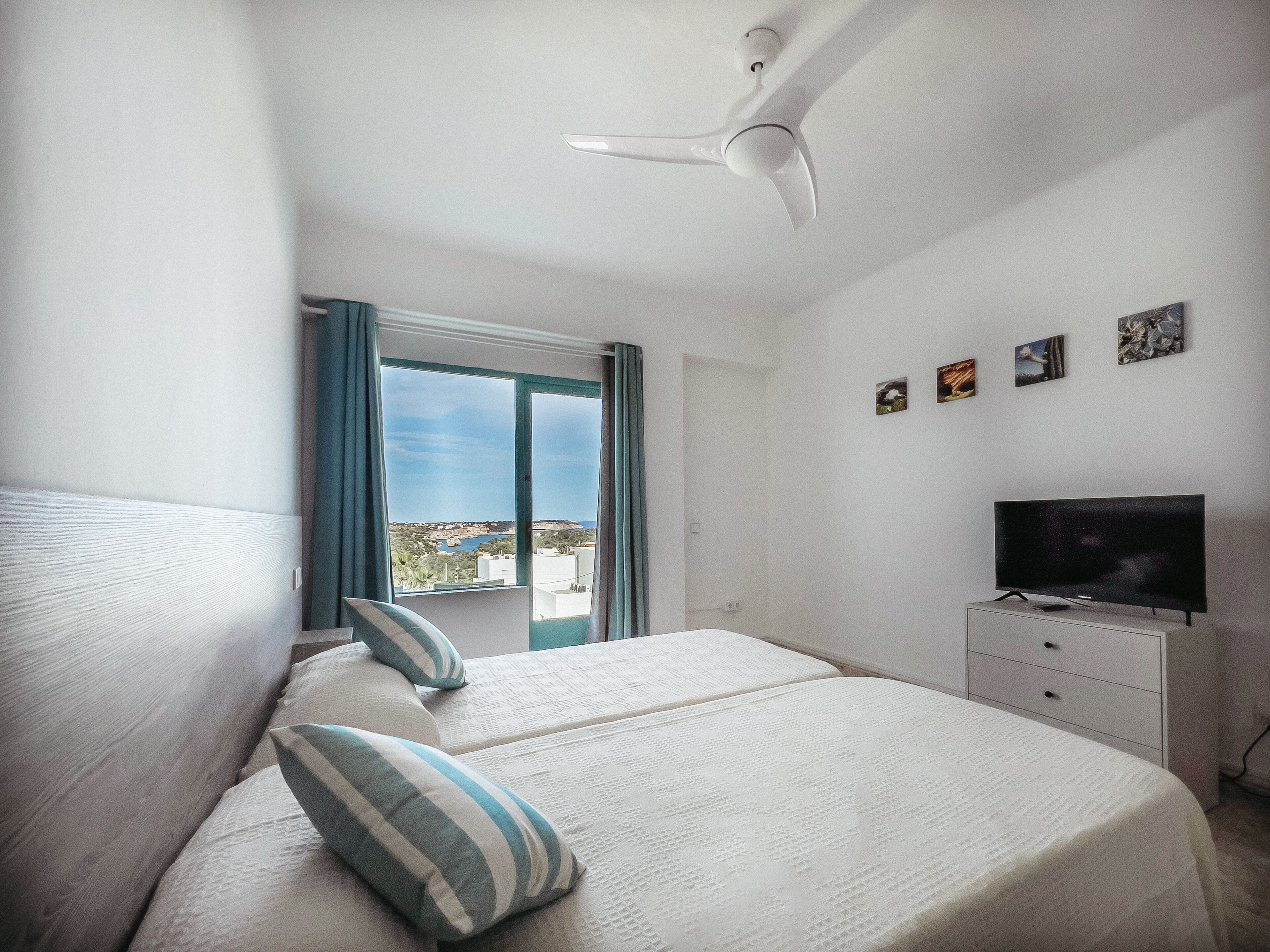 A bedroom with a bed, a TV, a ceiling fan, a window with curtains, and a balcony view.