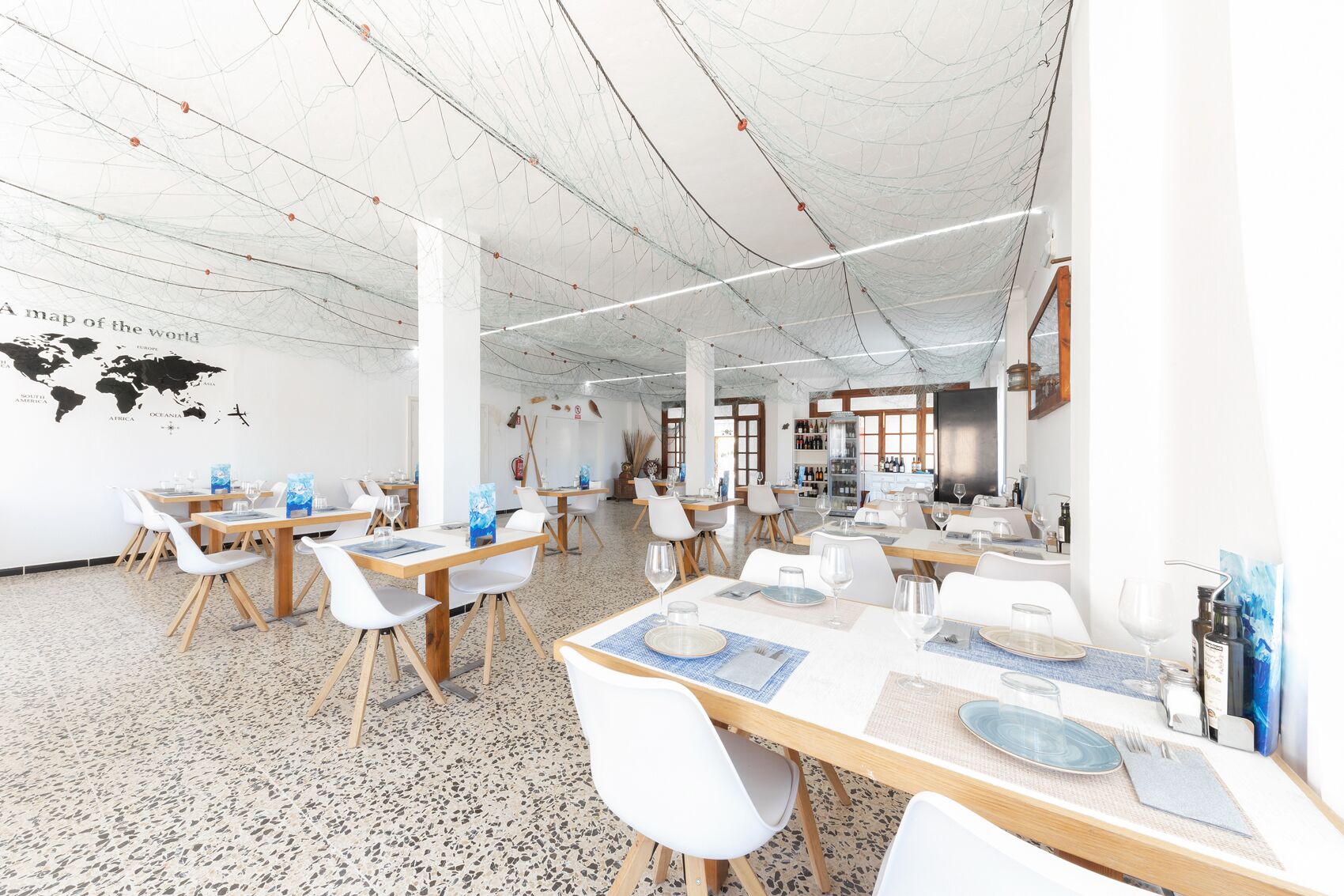 Un comedor amplio con techos altos y un mapa del mundo en la pared. Las mesas están dispuestas con sillas blancas y manteles individuales azules.